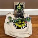 A soft cover EZD6 book standing up. In front is a tote with the EZD6 logo on the front, a set of green d6s with EZ where the 6 would, two pins, a magnet, and several stickers.
