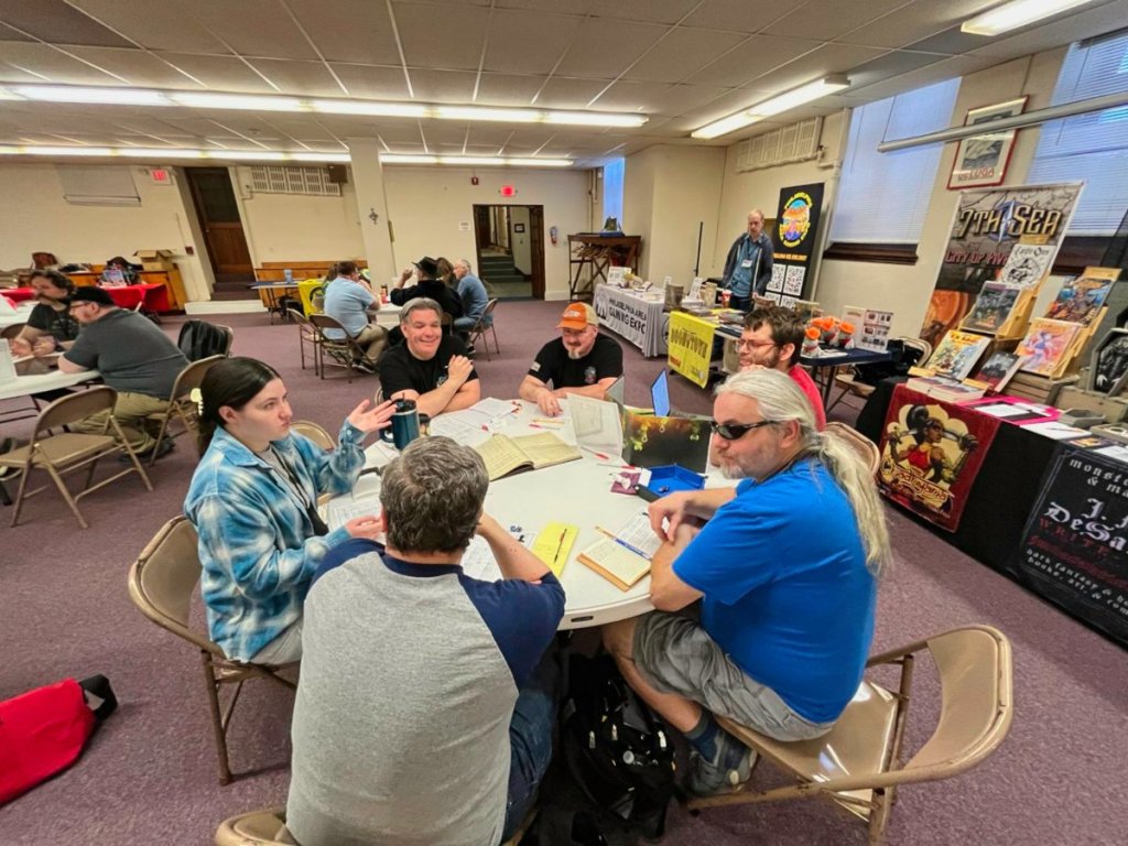 Players at a circular table discuss how to make their next move. Behind them are vendor tables.