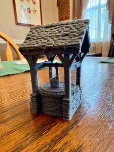 A 3d printed well, with a stone base and a slate roof. A bucket is placed on the well’s cover.