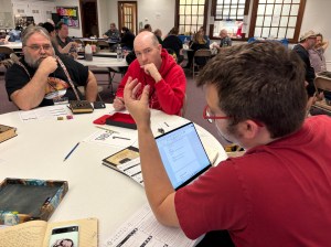 A game master describes a scene while glancing over his laptop. Two players look on in expectation